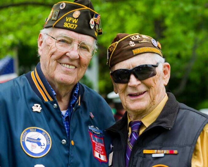 An elderly Korean War Veteran smiles at the camera with a fellow Veteran